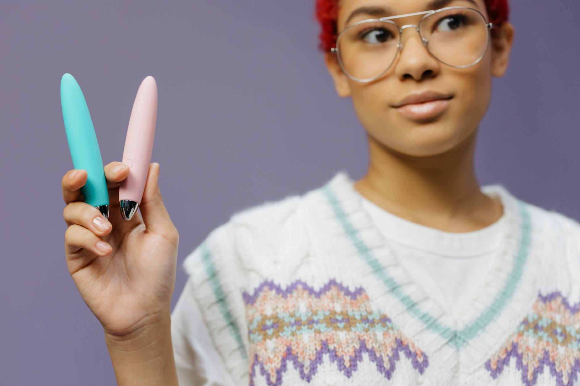 Une jeune femme portant des lunettes tenant deux vibromasseurs colorés contre un fond violet.