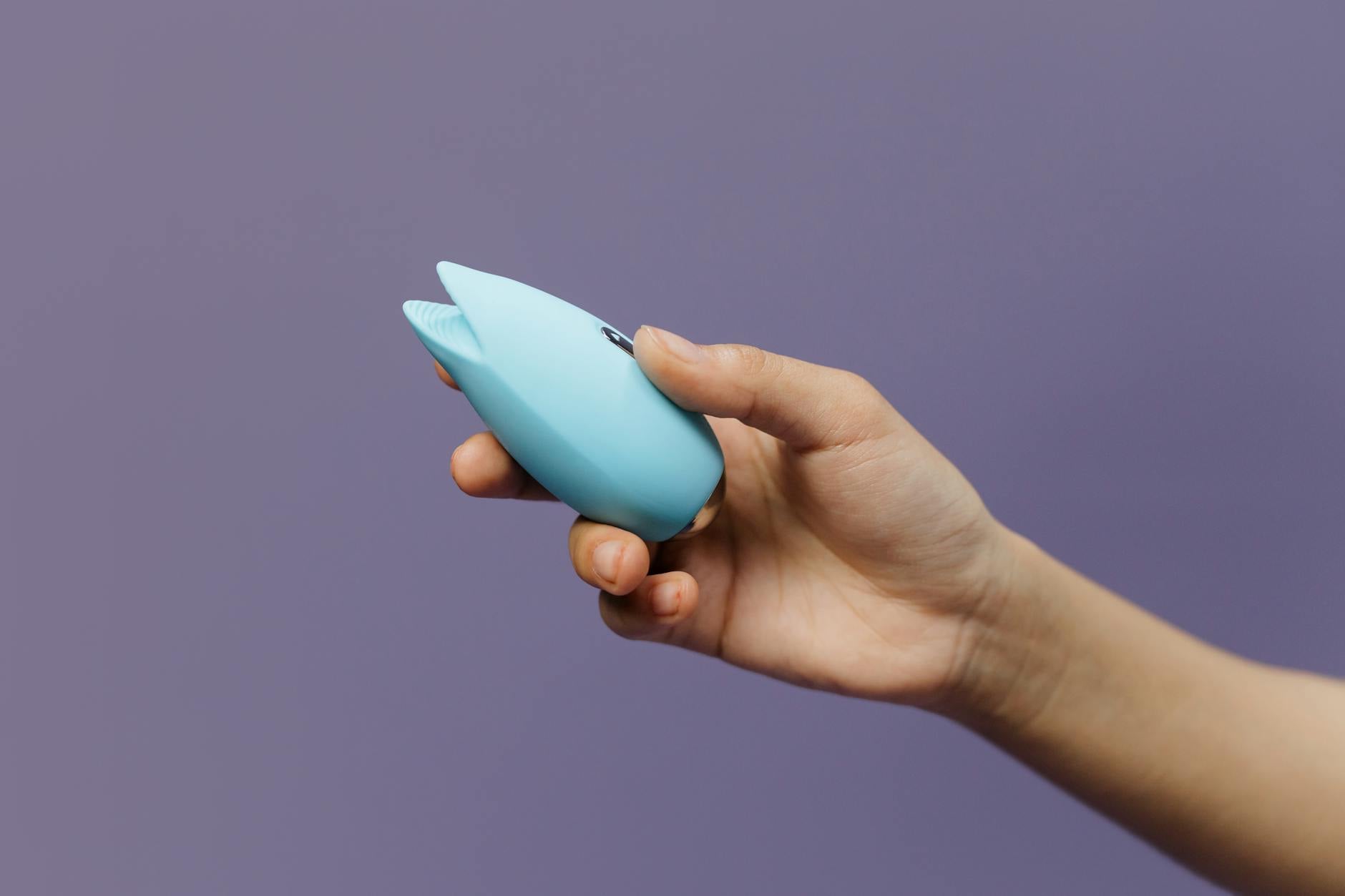 A hand holds a blue silicone vibrator against a purple background, representing self-pleasure and personal wellness
